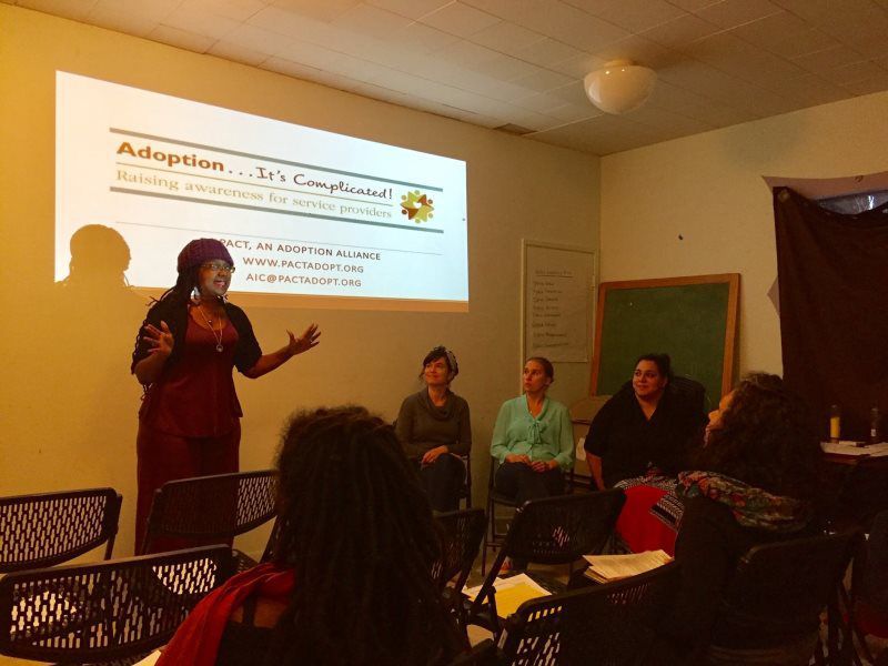Woman giving presentation about adoption. Audience members seated, four other women on stage.