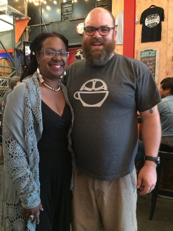 Woman in black dress and man in grey shirt posing together, smiling in a restaurant.