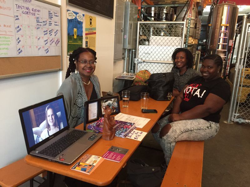 Three people sit at a table in a brewery. A laptop displays a woman's face, a whiteboard is in the background.