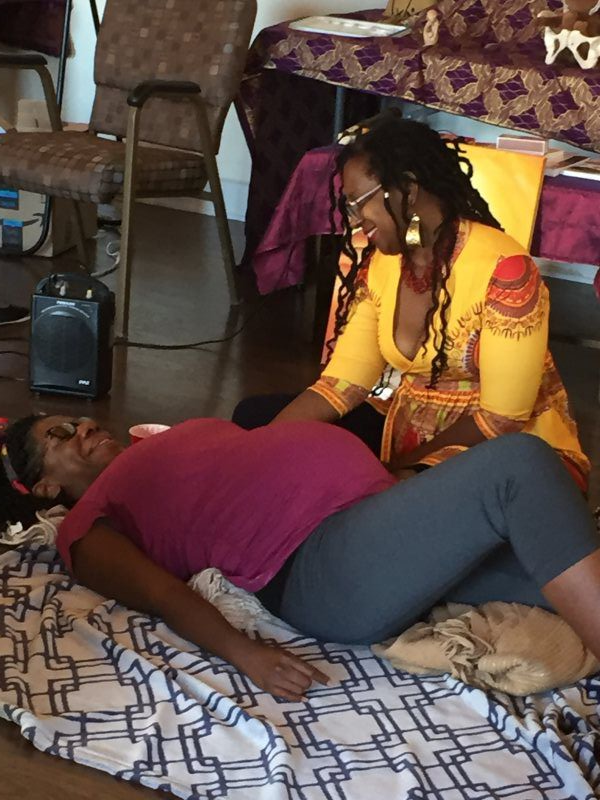 Pregnant person lying down receives care from a person with dreadlocks, both smiling. Indoors, on a patterned blanket.