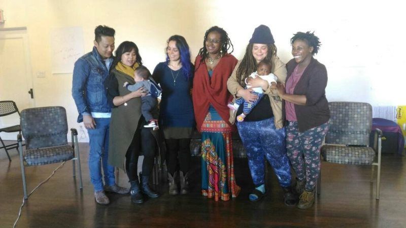 Group of people, some holding babies, posing indoors near chairs.