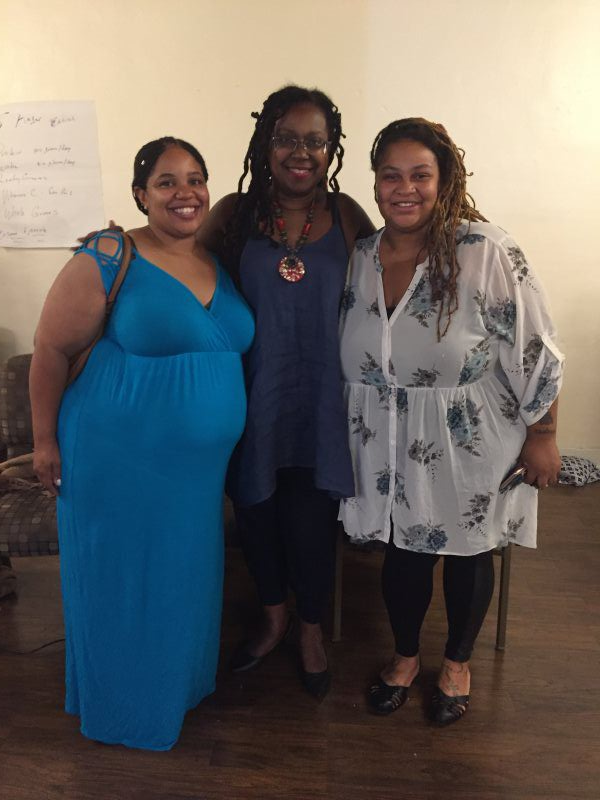 Three women smiling, posing for a photo.  One wears blue dress, another a blue top, and one a white top.