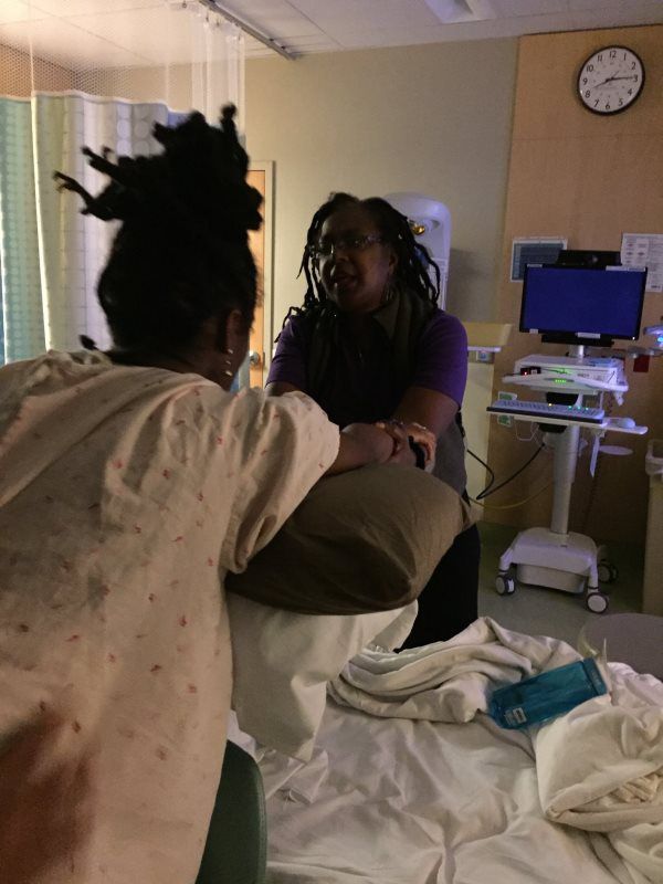 Two people talking near a hospital bed. One sits on the bed, back to camera, other sits beside, facing.