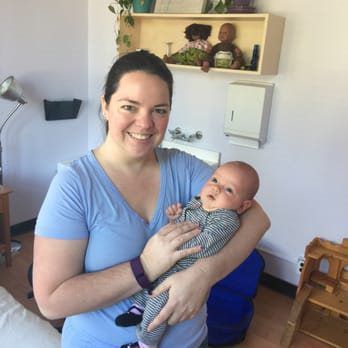 Woman in blue shirt holding baby, both smiling. Room with shelves, sink, and medical equipment.