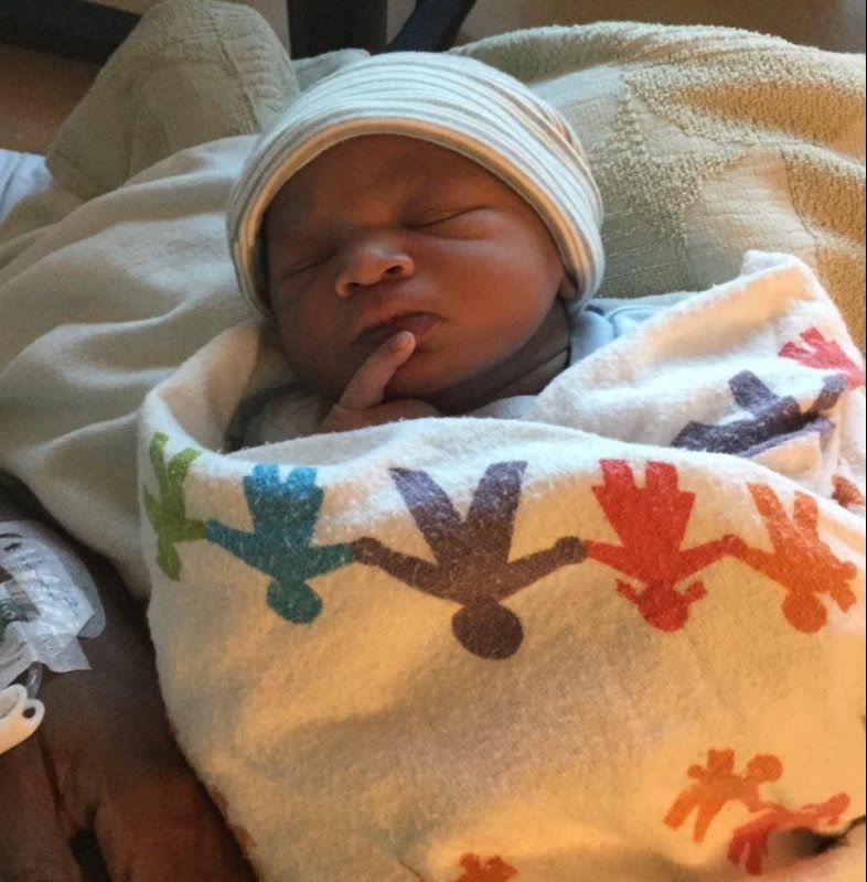 Newborn swaddled in blanket, wearing a hat, with finger on chin.