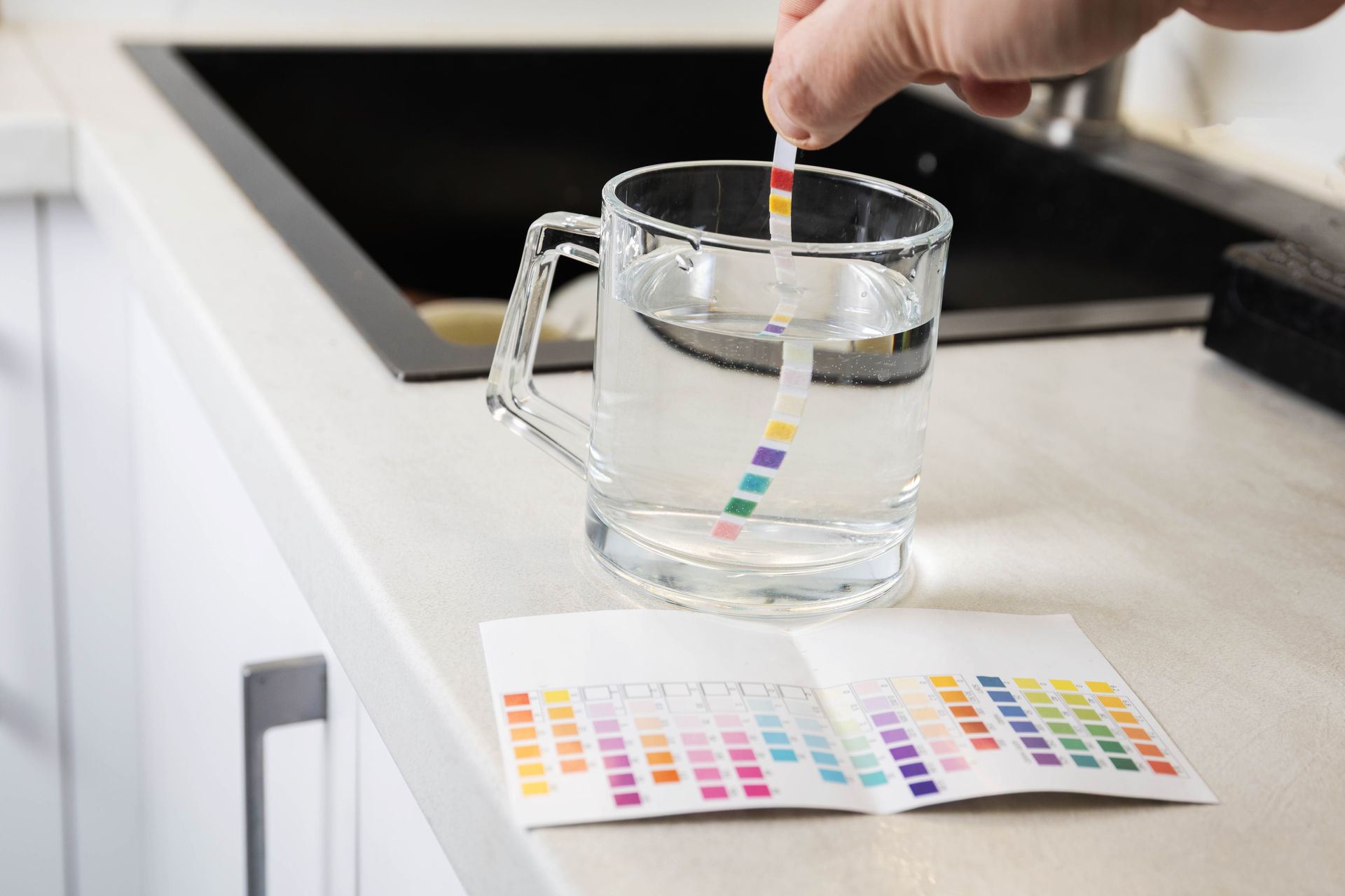 Hand holding a pH test strip above a glass mug of water beside a color chart on a kitchen counter