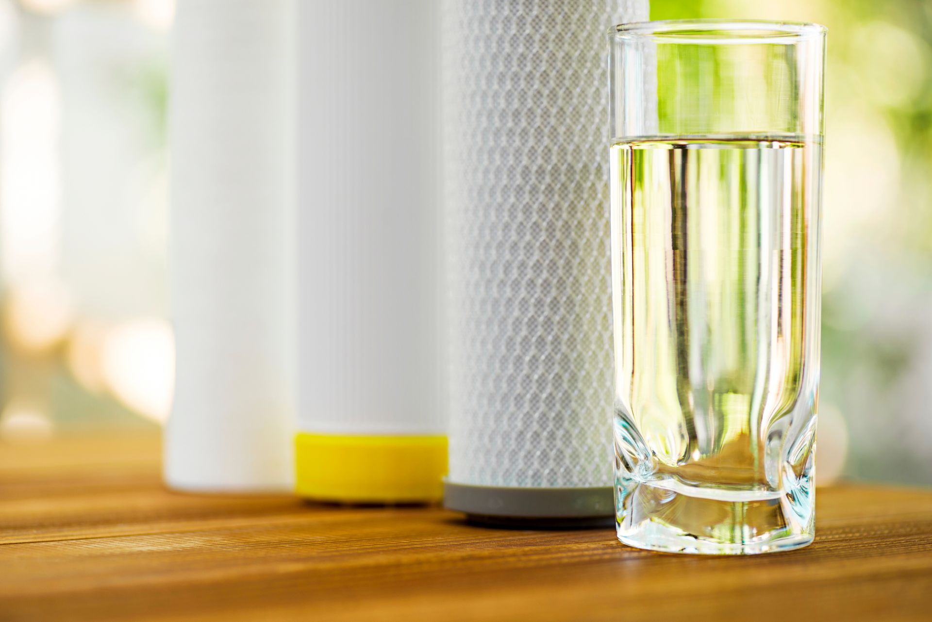 A glass of purified water and three filter cartridges on a wooden table with a natural background.
