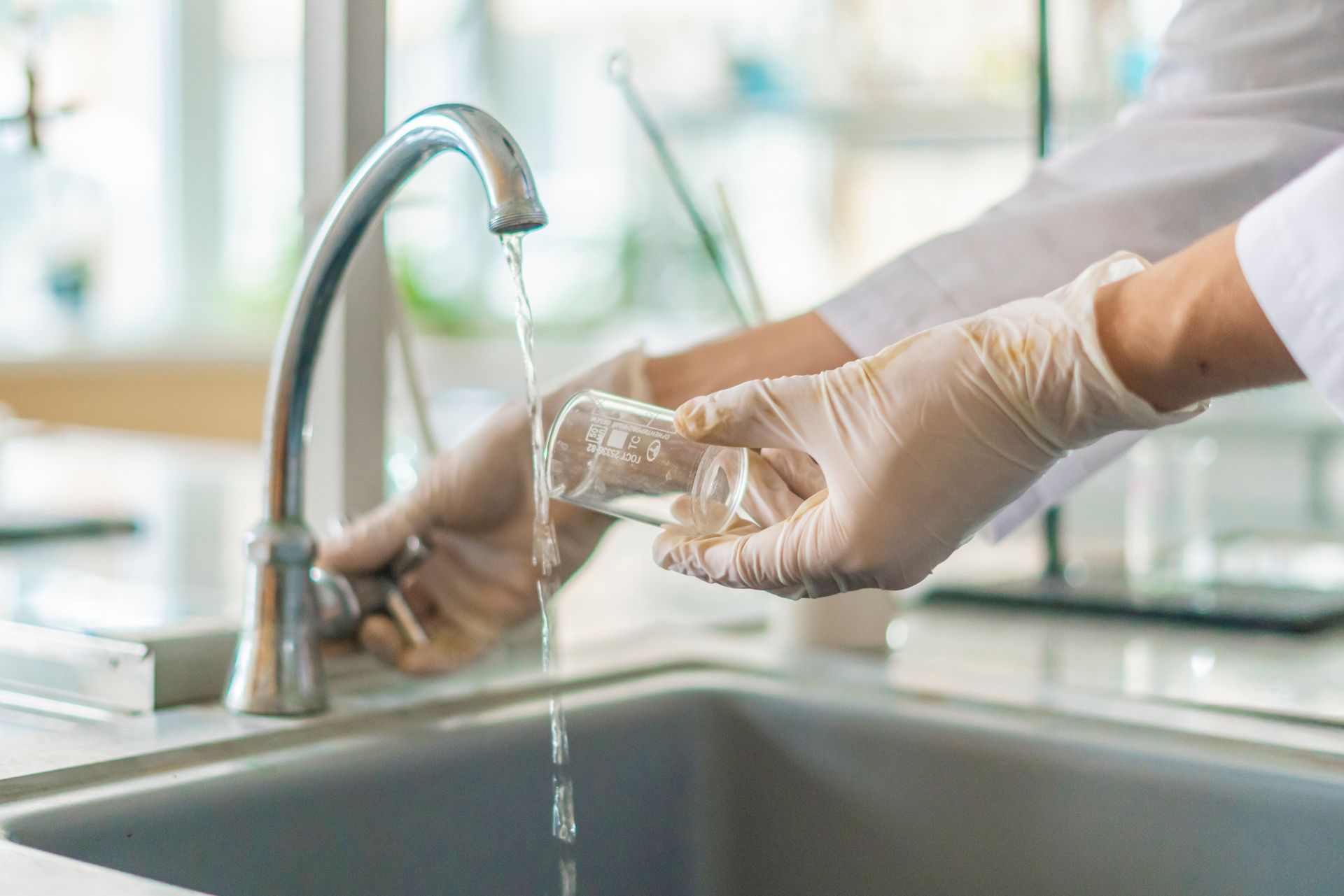 Gloved hands washing a clear glass under a running kitchen faucet