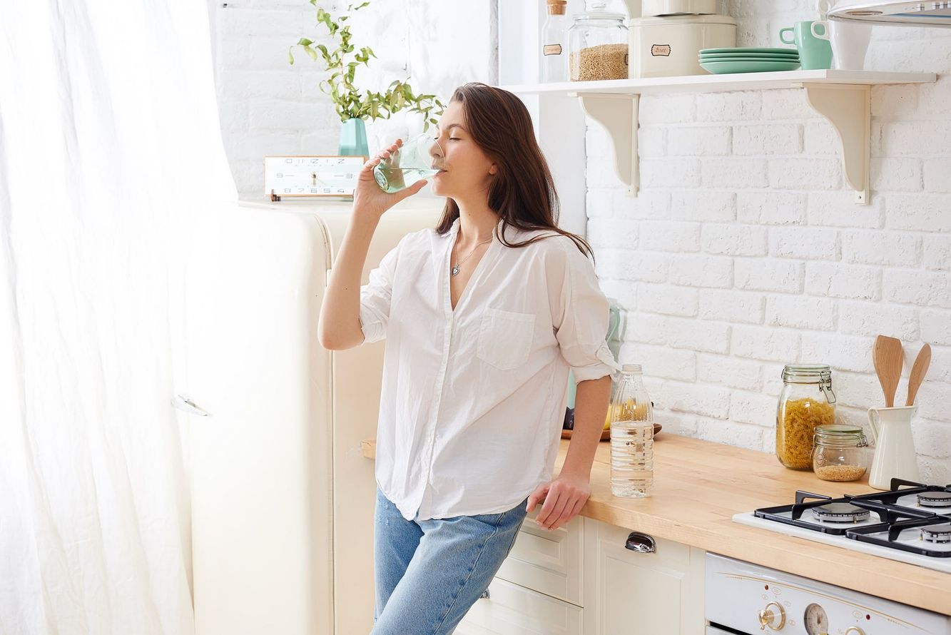 Woman in a bright kitchen drinking from a glass, standing by a white brick wall and wooden counter. Woman in a bright kitchen drinking from a glass, standing by a white brick wall and wooden counter.