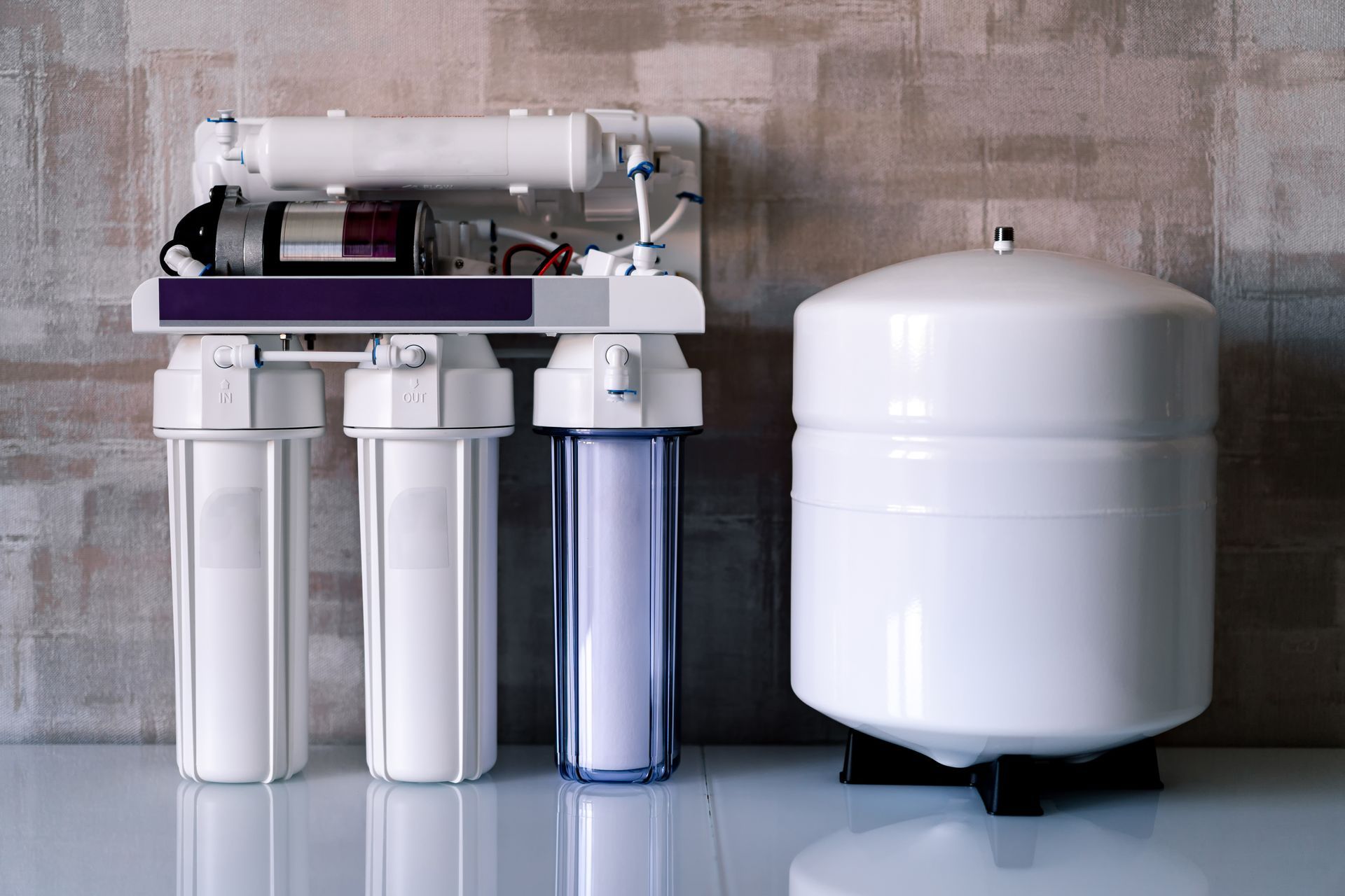 Water filtration system with three clear cartridges and a large white tank against a concrete wall