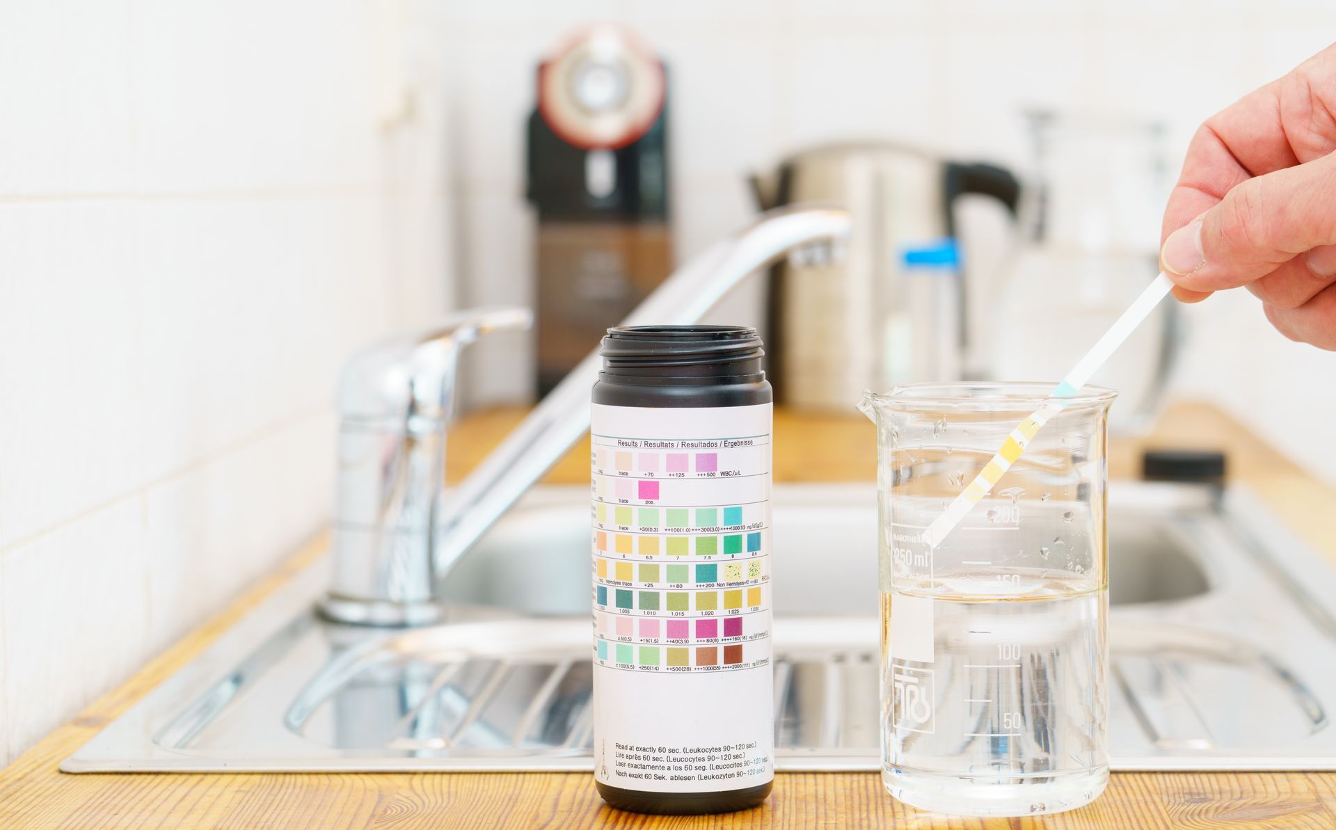 Hand holding a dropper over a glass of clear water beside a test container in a kitchen sink