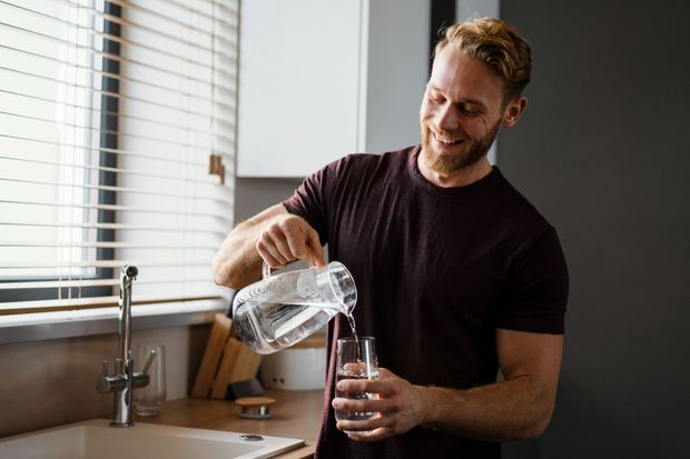 Man pouring water from a pitcher into a glass in a kitchen by the sink Man pouring water from a pitcher into a glass in a kitchen by the sink