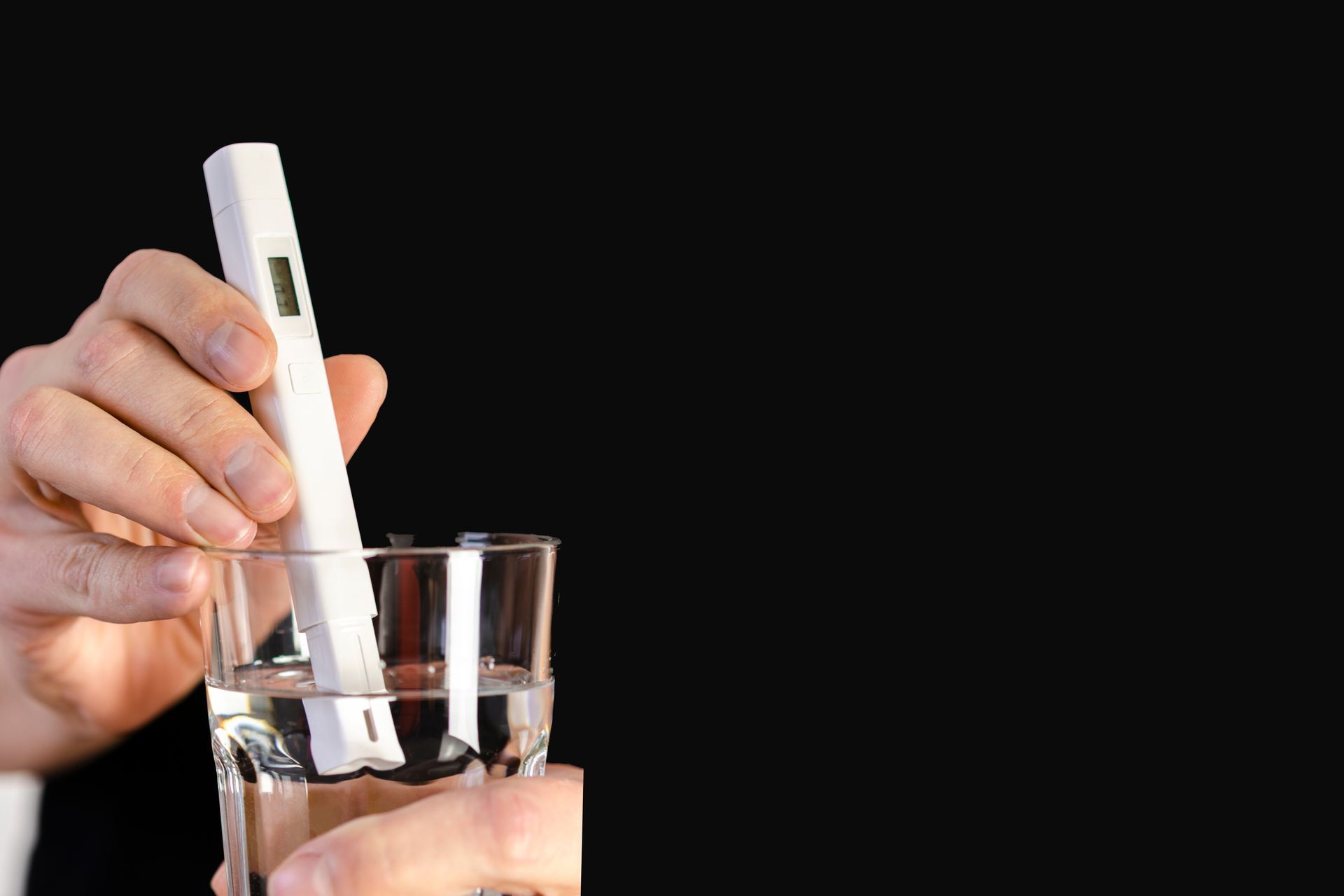 Hand holding a white digital thermometer over a glass of water on a black background Hand holding a white digital thermometer over a glass of water on a black background