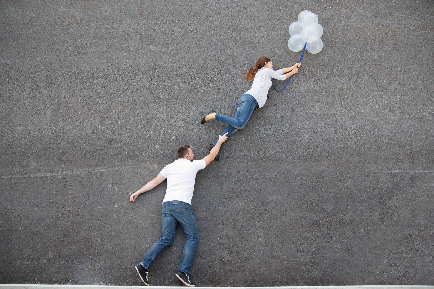 Ein Mann hält die Hand einer Frau, die mit weißen Luftballons vor einer grauen Wand davonschwebt.
