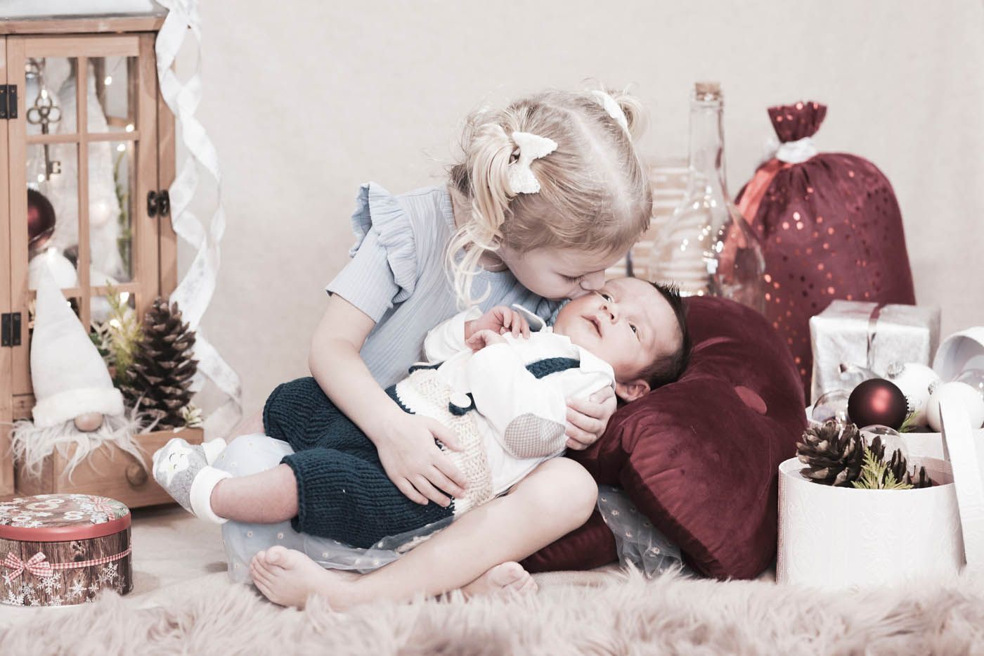 Ein Mädchen küsst die Stirn eines Babys. Sie sitzen inmitten von Weihnachtsdekorationen.