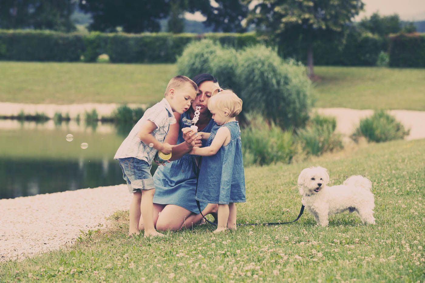 Eine Frau und zwei Kinder pusten Seifenblasen an einem See mit einem kleinen weißen Hund.