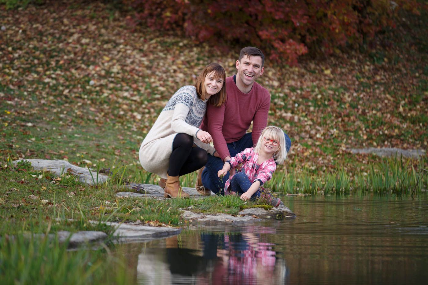 Eine lächelnde Familie an einem Teich. Eine Frau hockt, ein Mann steht. Ein Mädchen in einem rosa T-Shirt spielt am Wasser.