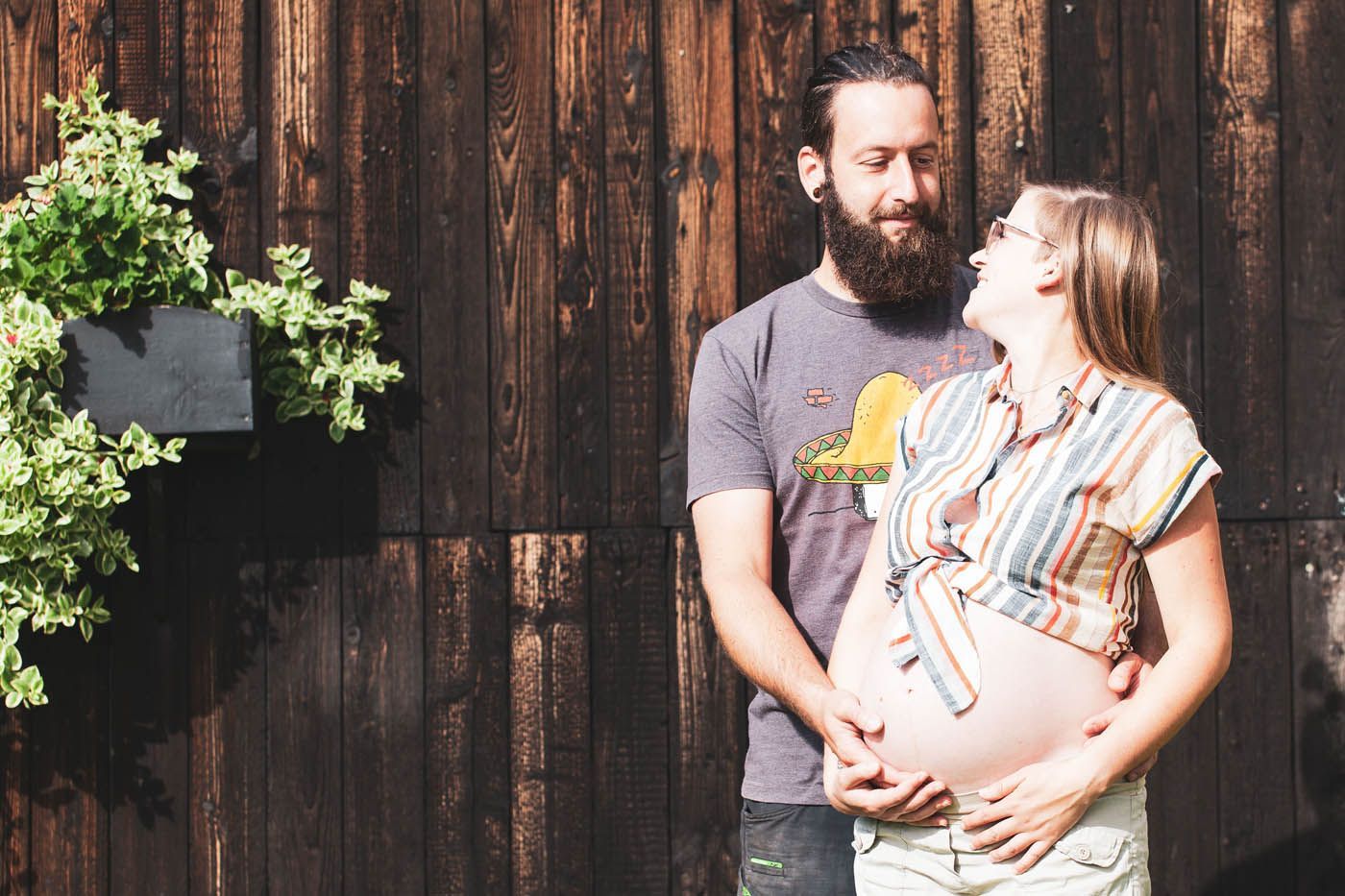 Schwangere Frau und ihr Partner umarmen sich lächelnd. Sie stehen draußen an einer Holzmauer mit viel Grün.