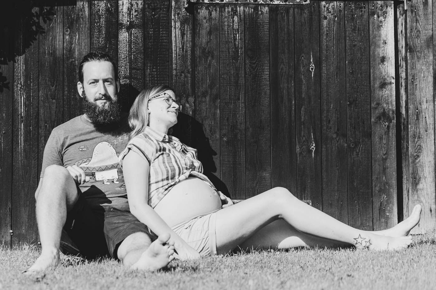 Ein Mann mit Bart und eine schwangere Frau sitzen lächelnd auf einer Wiese, lehnen an einer Holzwand.