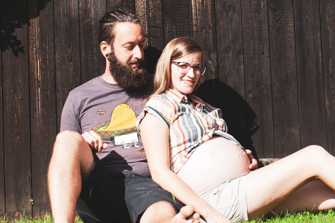 Schwangere und ihr Partner sitzen lächelnd auf einer Rasenfläche, an einen Holzzaun gelehnt.