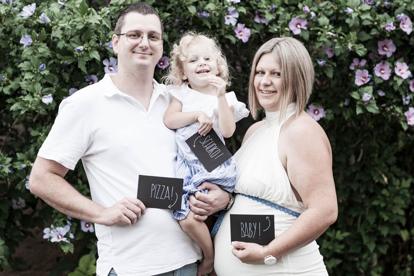 Dreiköpfige Familie hält Schilder; die Mutter ist schwanger und steht vor einem Busch mit lila Blüten.