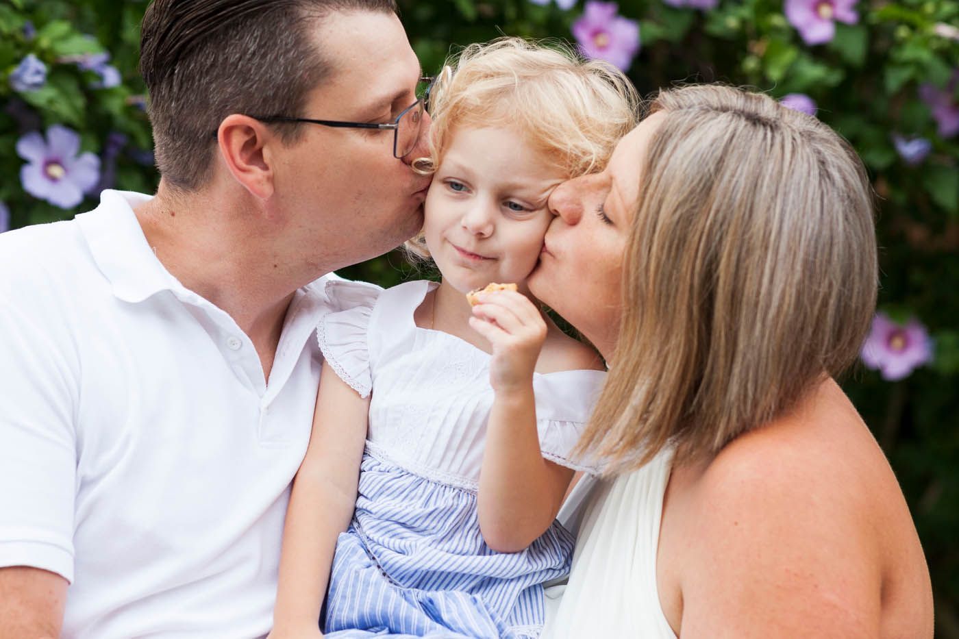 Dreiköpfige Familie, Eltern küssen die Wangen eines Kindes. Hintergrund mit Blumen.
