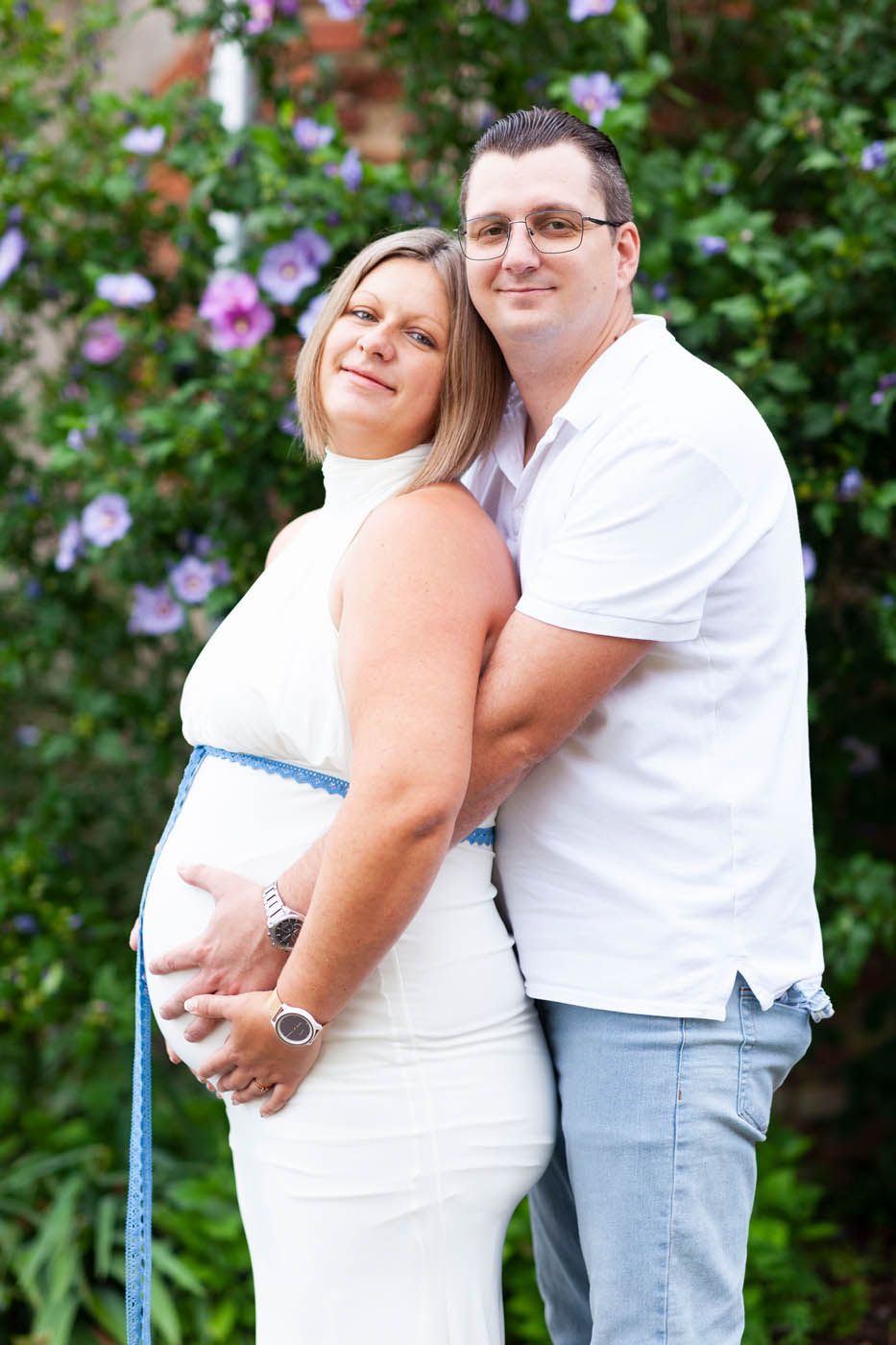 Schwangere Frau in weißem Kleid, umarmt von einem Mann in weißem Hemd, stehend neben blühenden Büschen.