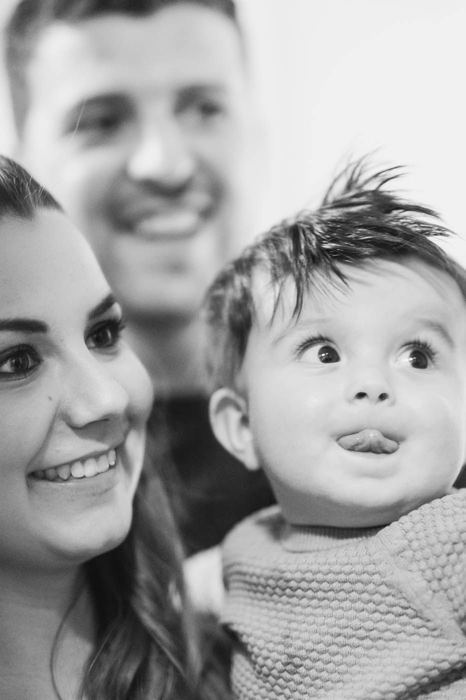 Familienporträt: Frau lächelt neben einem Kind, das die Zunge herausstreckt; im Hintergrund lächelt ein Mann.