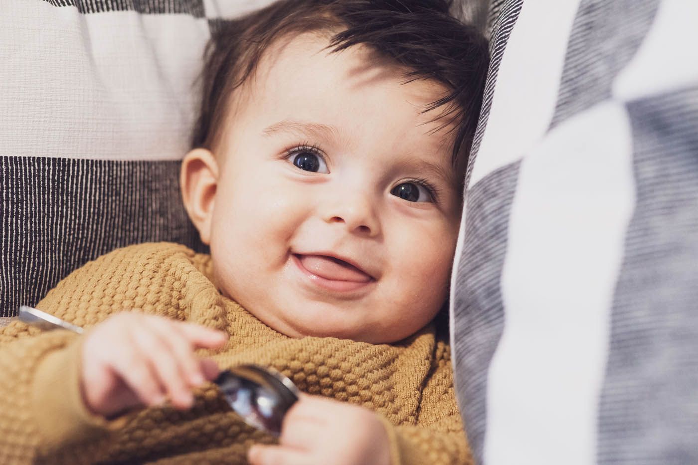 Ein lächelndes Baby in gelbem Pullover hält einen Löffel und liegt eingebettet in gestreiften Kissen.