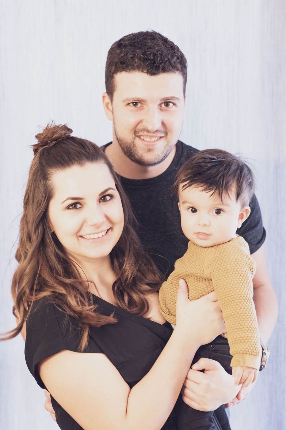 Dreiköpfige Familie lächelt in die Kamera: Frau hält ein Baby, Mann steht hinter ihnen.