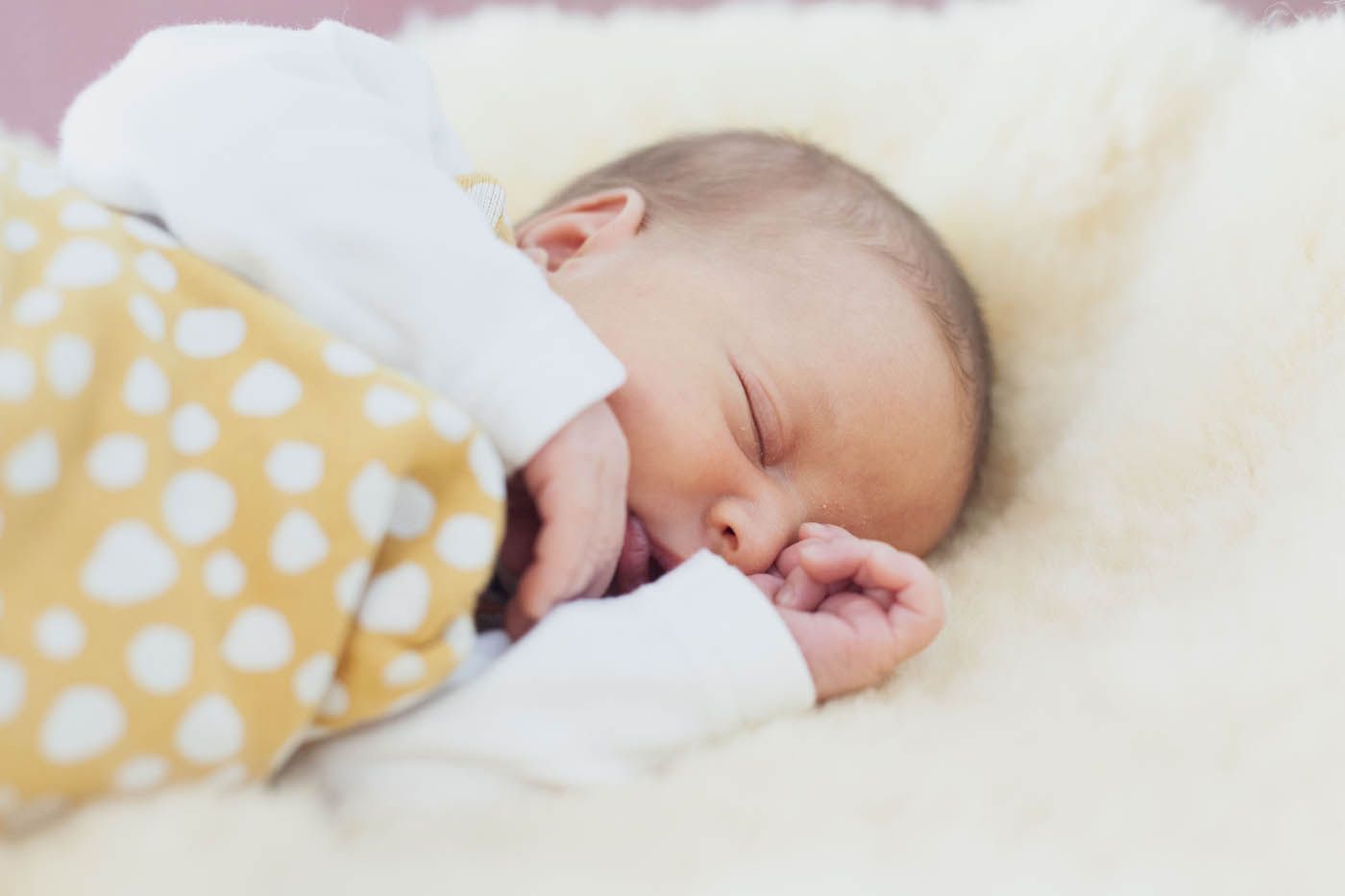 Ein schlafendes Baby liegt auf einer flauschigen weißen Unterlage, eingewickelt in eine gelb gemusterte Decke.