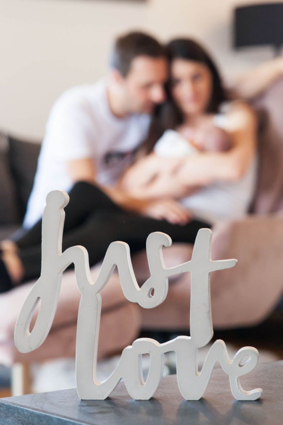 Weißes Schild mit der Aufschrift „Erste Liebe“; verschwommenes Bild eines Paares mit Baby im Wohnzimmer.