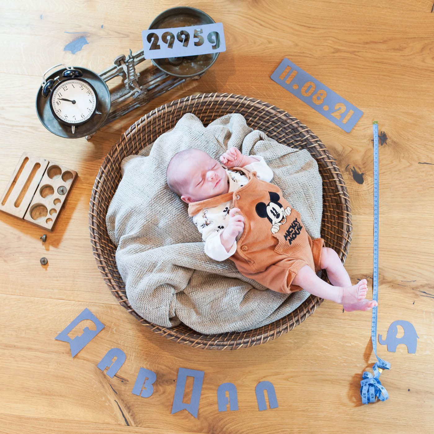 Neugeborenes Baby in orangefarbenem Mickey-Mouse-Outfit in einem Weidenkorb mit Gewichts- und Datumsmarkierungen.