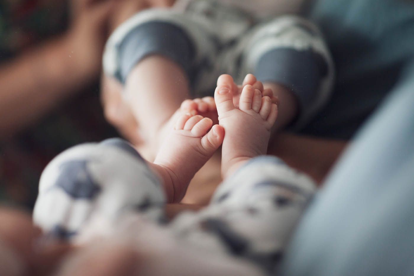 Nahaufnahme von Babyfüßen in hellblau-weiß gemusterten Hosen, die von den Händen eines Erwachsenen gehalten werden.