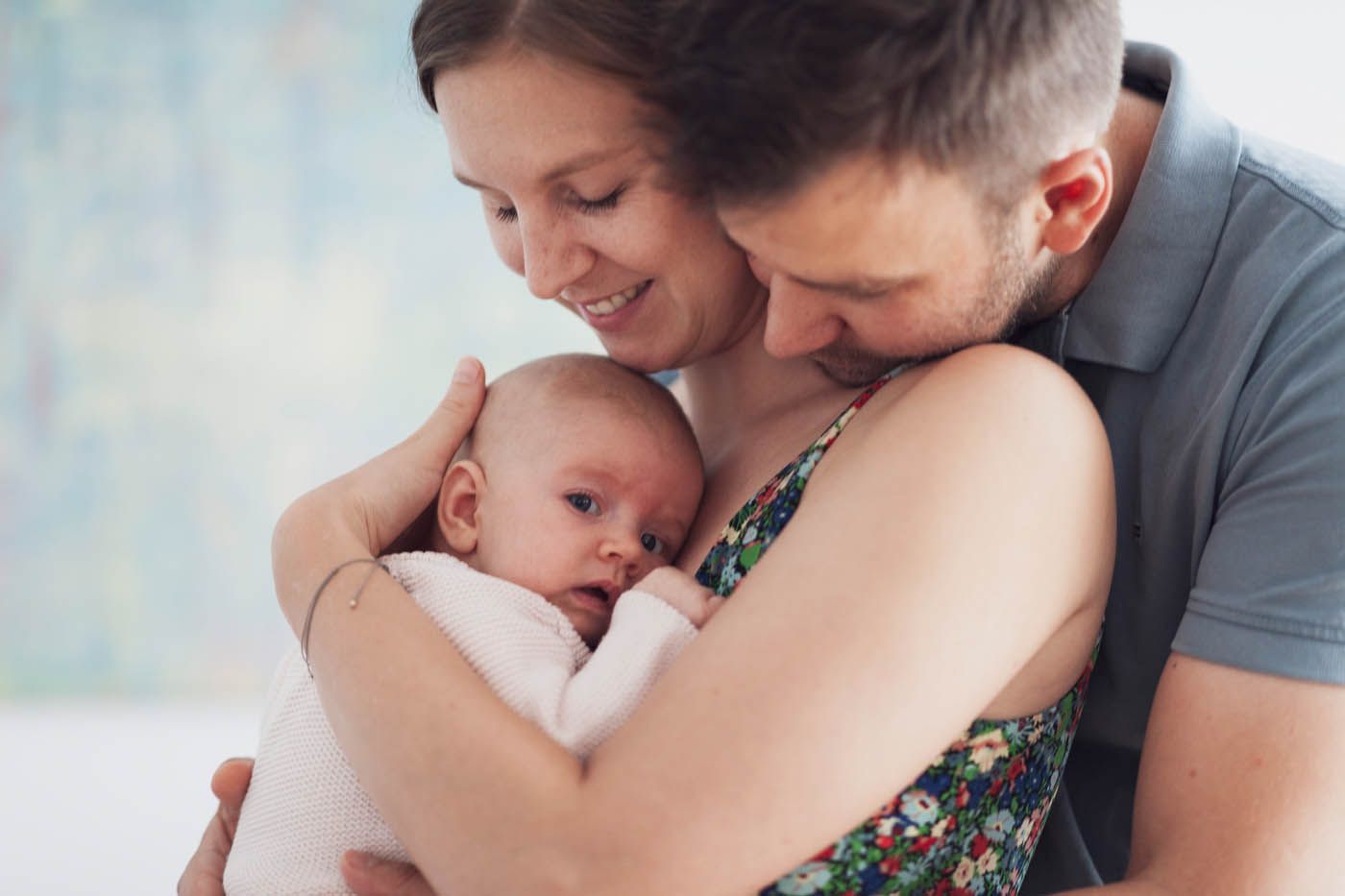 Eltern halten ein Baby. Die Frau wiegt das Kind im Arm, der Mann küsst ihre Schulter. Heller Hintergrund.