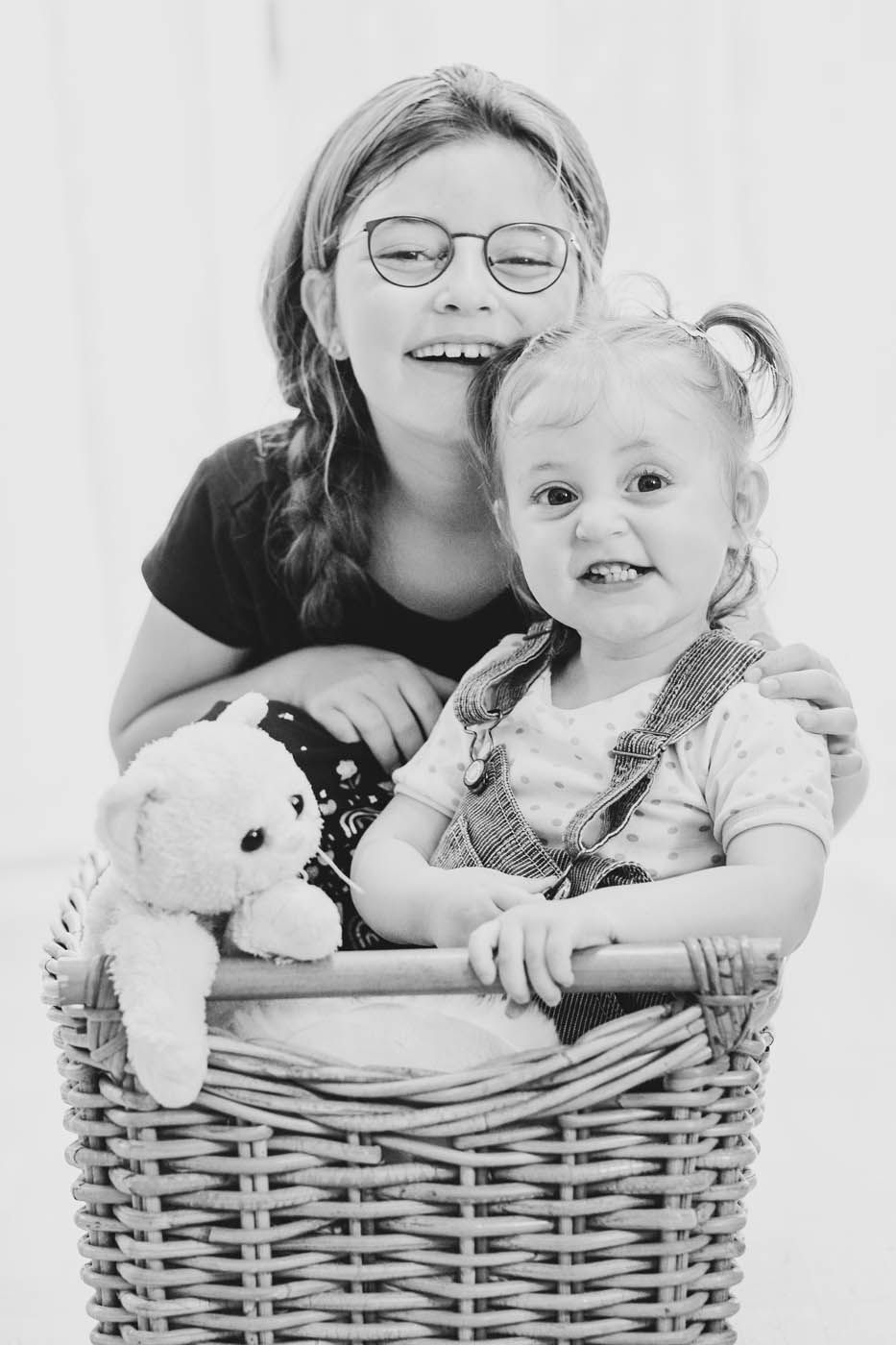 Mädchen mit Brille umarmt Kleinkind in einem Weidenkorb, beide lächeln. Teddybär im Korb.
