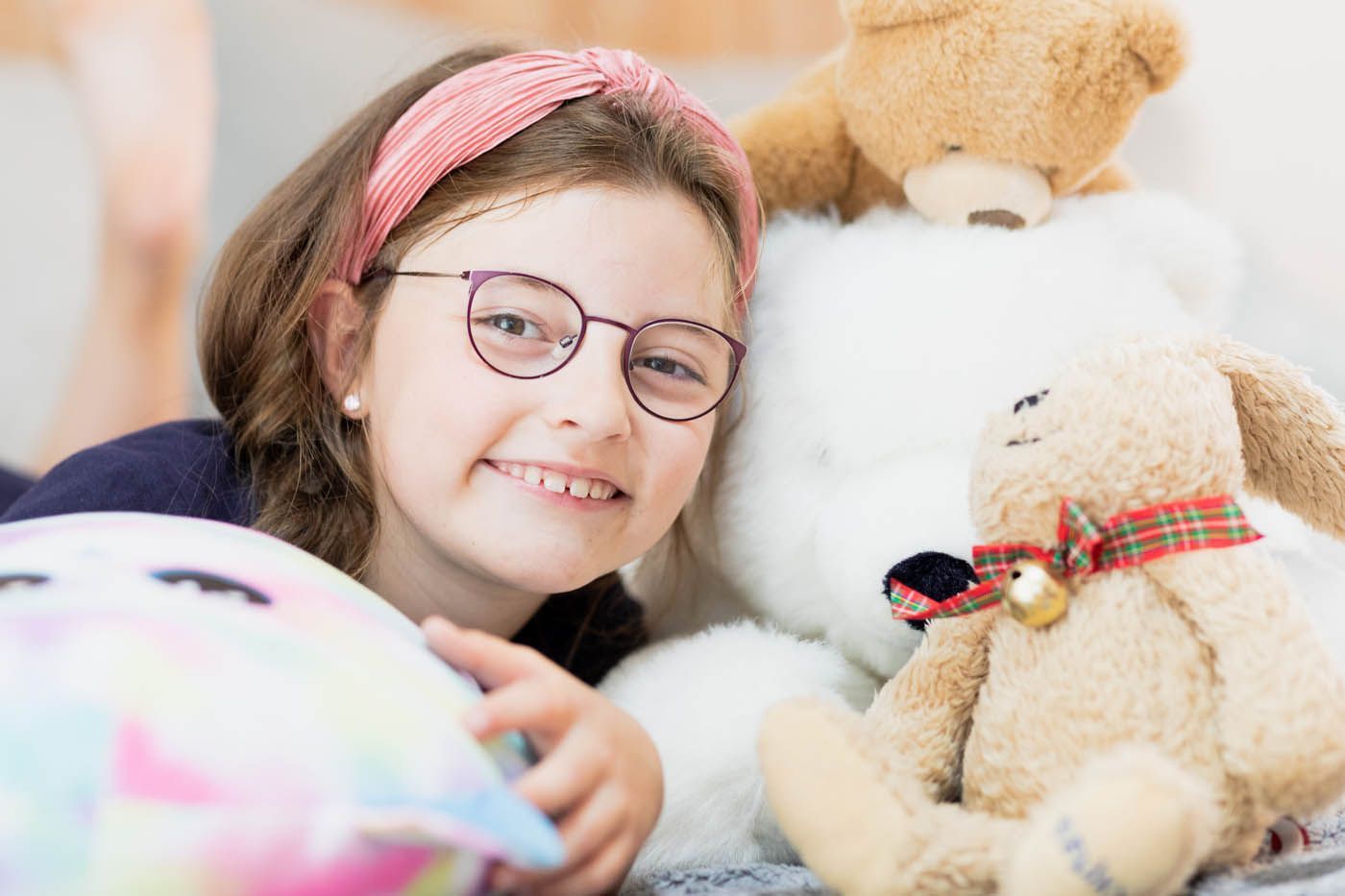 Mädchen mit Brille lächelt, umgeben von Stofftieren und einem Kissen.