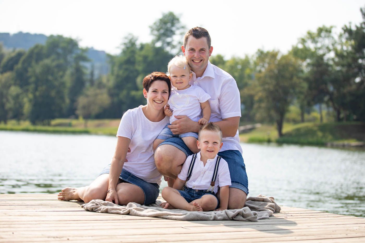 Eine vierköpfige Familie sitzt lächelnd auf einem Holzsteg an einem See und trägt weiße Hemden und Jeansshorts.