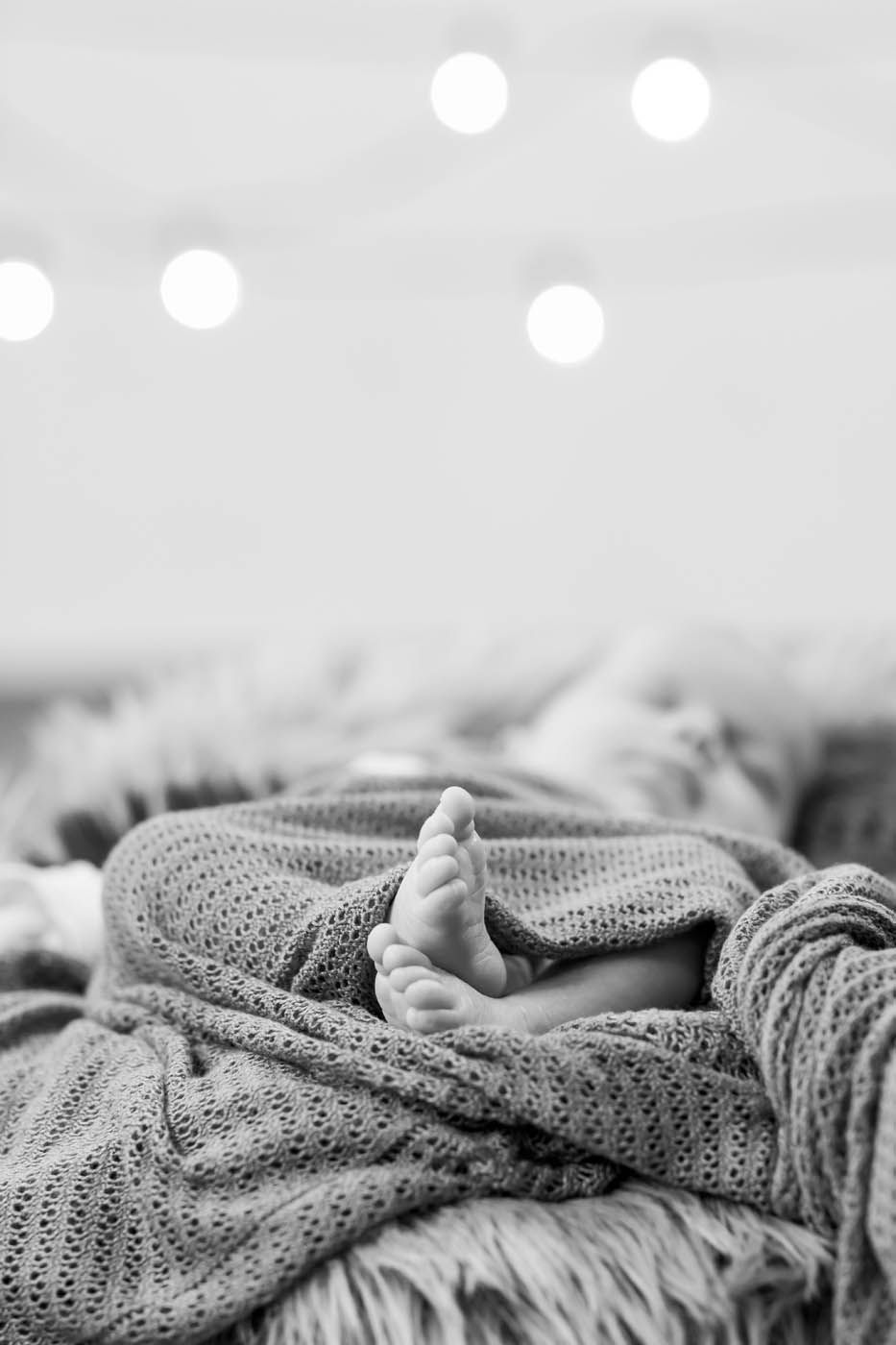 Babys Füße lugen unter einer grauen Strickdecke auf einer flauschigen Unterlage hervor, im Hintergrund sanftes Licht.