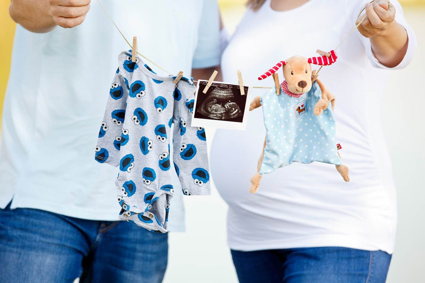 Schwangere Frau mit Wäscheleine, an der Babykleidung und ein Ultraschallbild hängen; Partner daneben.