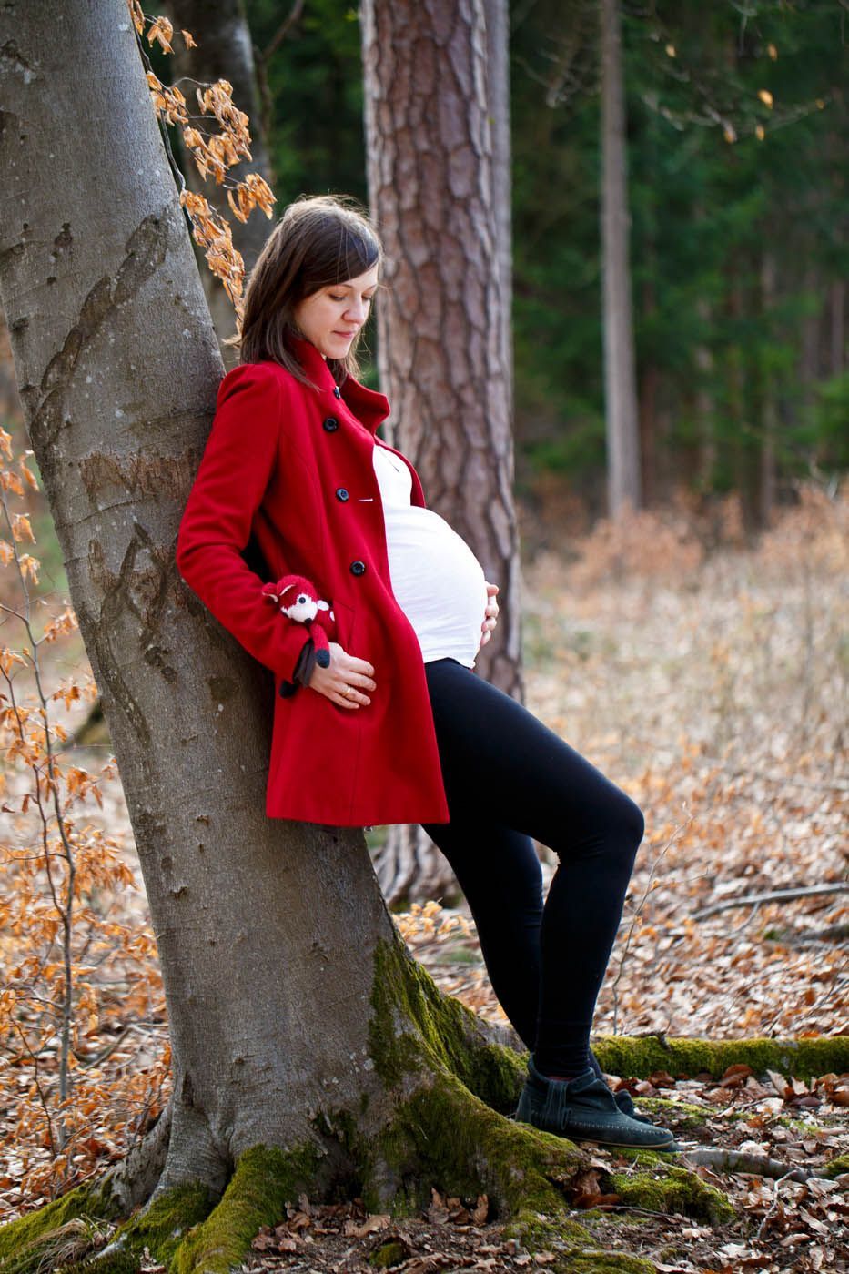 Schwangere Frau in rotem Mantel, die sich in einem Wald an einen Baum lehnt.