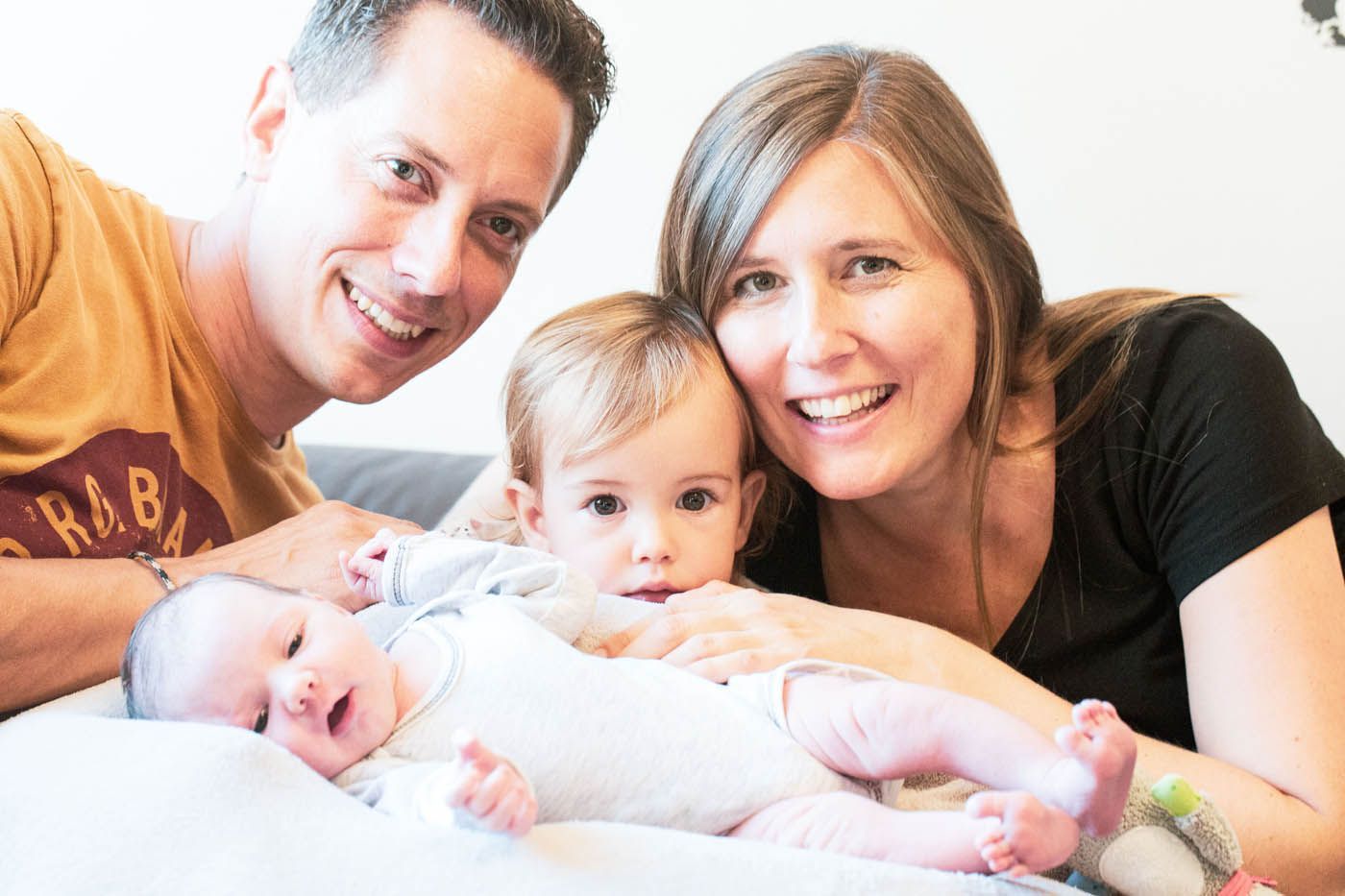 Eine vierköpfige Familie lächelt in die Kamera: zwei Eltern und zwei Babys liegen auf einer weißen Decke.