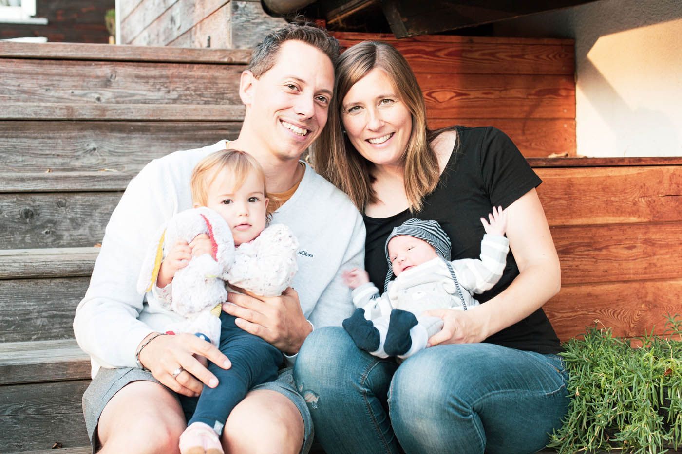 Eine vierköpfige Familie sitzt lächelnd auf einer Holztreppe. Zwei Erwachsene halten kleine Kinder im Arm.