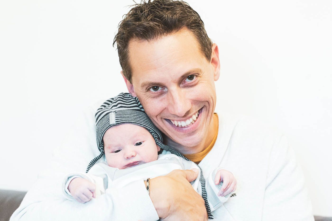 Mann lächelt und hält ein Baby mit gestreifter Mütze im Arm. Heller Hintergrund.