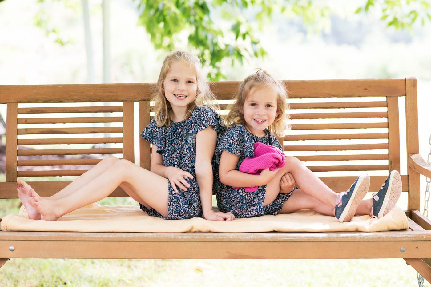 Zwei junge Mädchen lächeln auf einer Holzbank, beide tragen die gleichen geblümten Kleider.