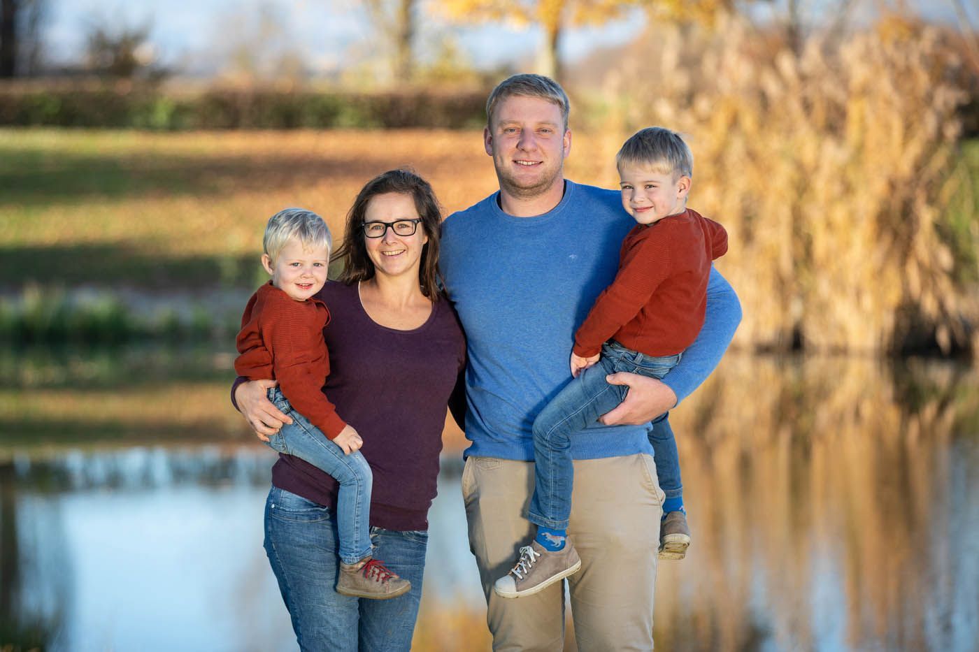 Eine vierköpfige Familie lächelt an einem See: Eltern mit kleinen Kindern, alle in legerer Kleidung. Herbstliche Szenerie.