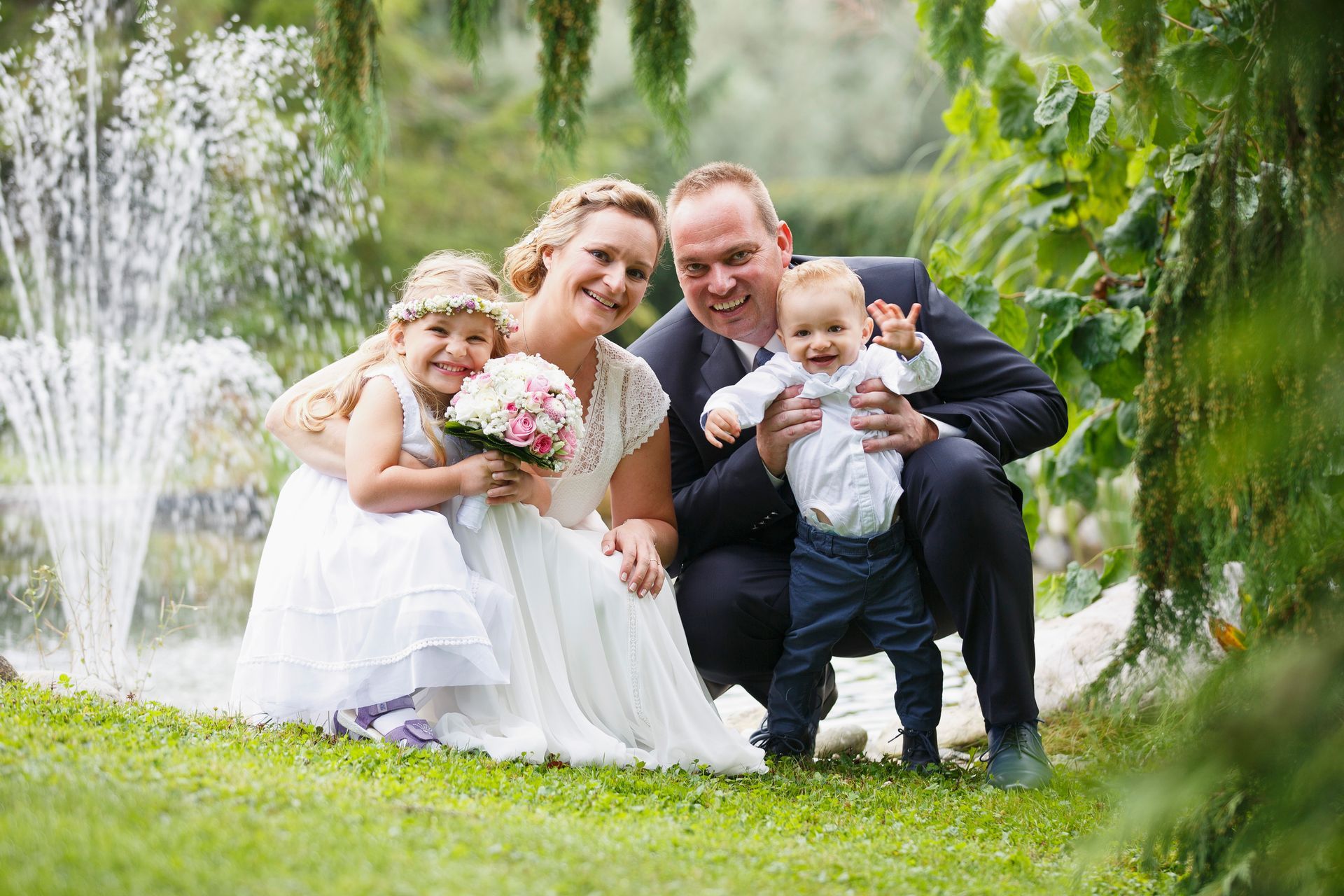 Eine vierköpfige Familie lächelt an einem Brunnen; die Eltern tragen Hochzeitskleidung.