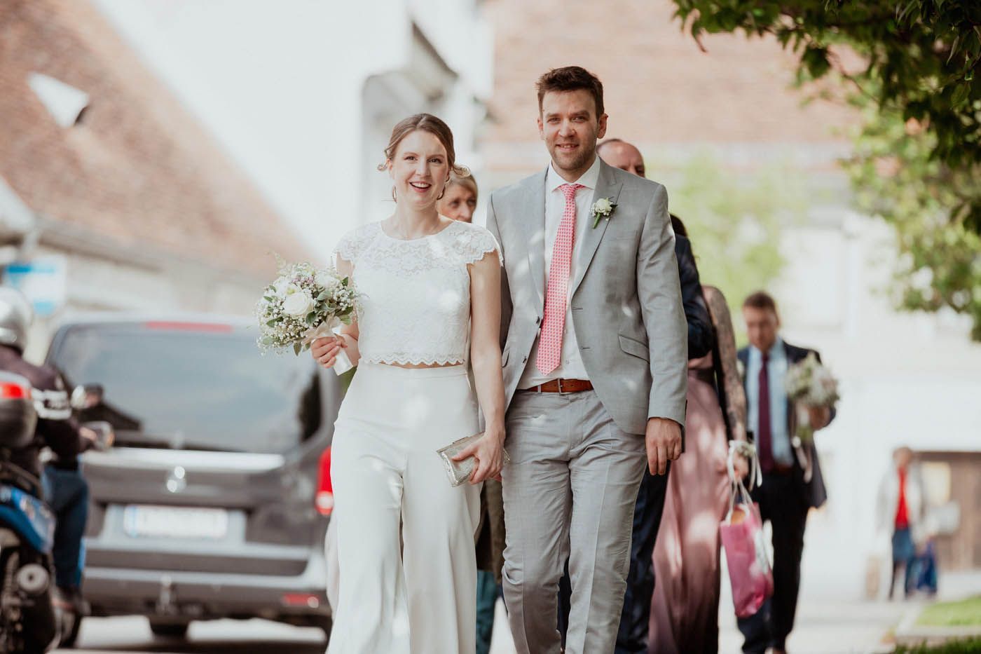 Braut und Bräutigam spazieren Hand in Hand mit den Hochzeitsgästen. Draußen in einer Stadt. Der Mann trägt einen grauen Anzug, die Frau einen weißen Hosenanzug.
