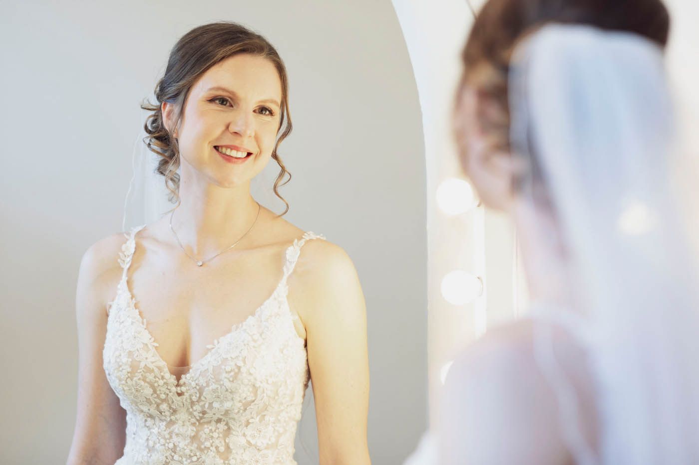 Die Braut im Hochzeitskleid blickt mit einem sanften Lächeln in den Spiegel. Schleier und hochgesteckte Frisur.