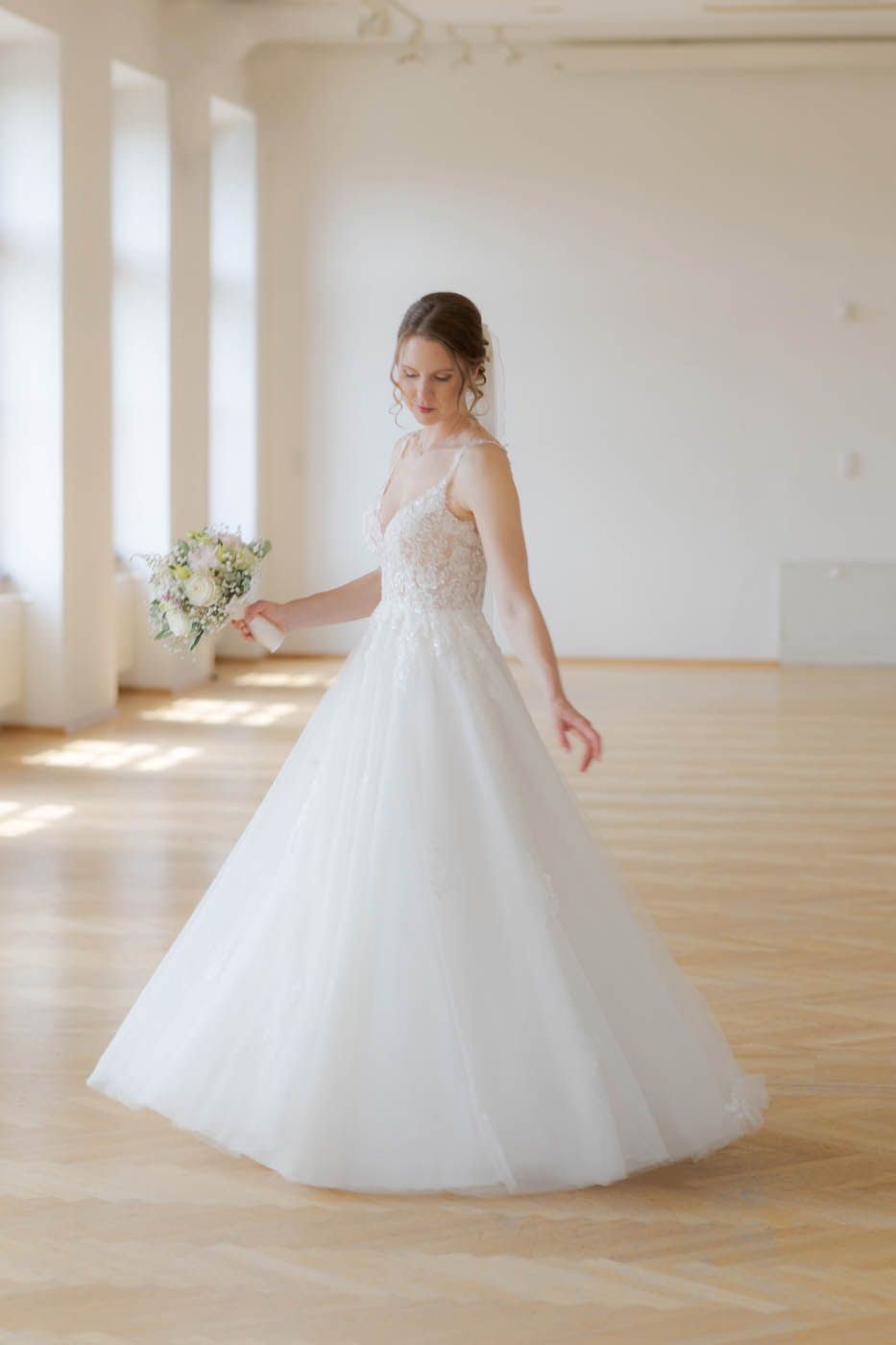 Eine Braut im weißen Hochzeitskleid hält Blumen in den Händen und steht in einem sonnendurchfluteten Raum.